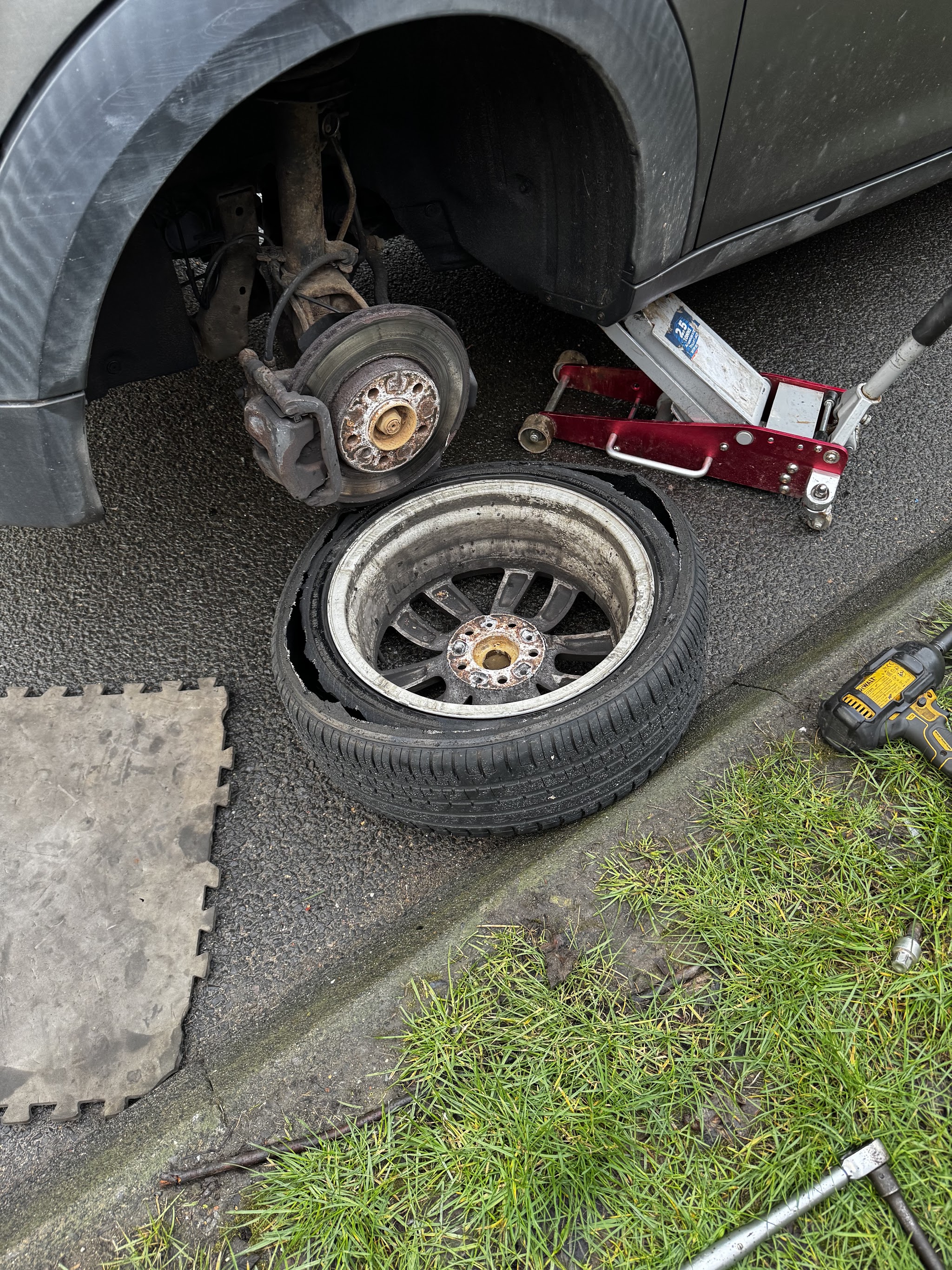 A shredded blown tyre on its alloy wheel beside a car on a trolley jack at the kerb, with an impact gun and kneeling mat